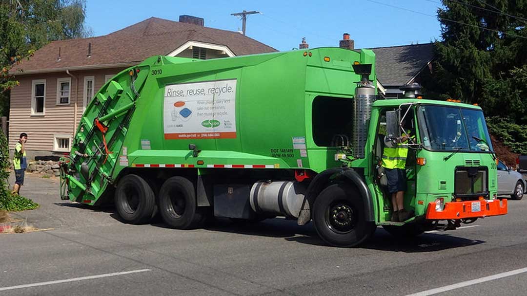 Large vinyl banner attached to side of waste disposal truck reading 'Rinse, Reuse, Recycle.' with illustration of plastic containers.