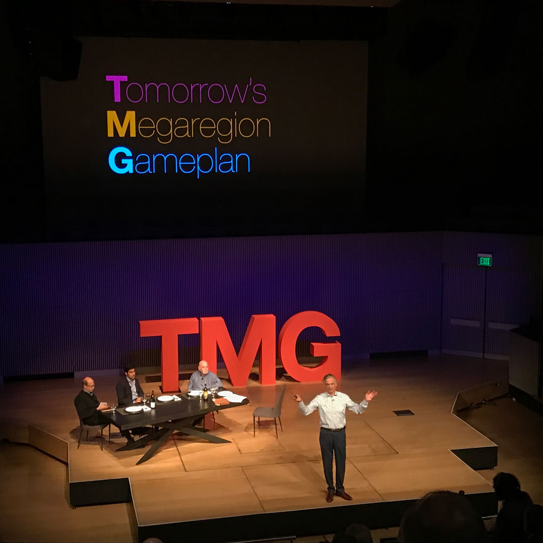 TMG Chairman standing center stage delivering closing remarks with large red TMG letters behind him, and a projected screen above which reads 'Tomorrows Megaregion Goals'.