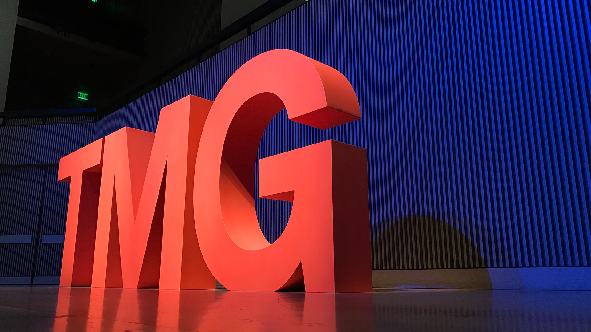 5 foot-tall wooden letters 'TMG' painted bright red on stage with blue light background.