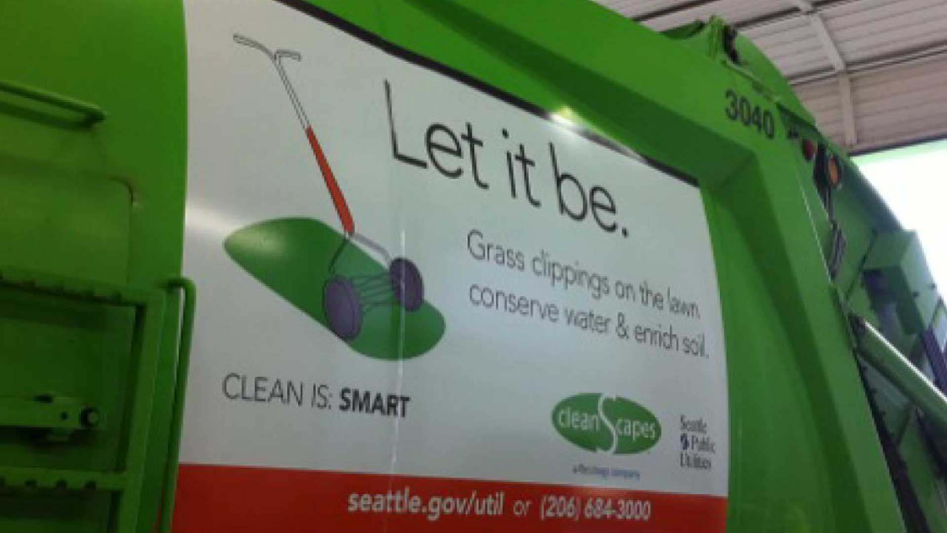 Large vinyl banner attached to side of waste disposal truck reading 'Let it be.' with illustration of manual grass mower and grass clippings.